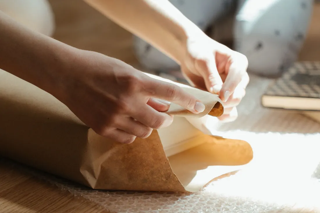Hände schlagen ein kleines Gefäß sorgfältig in braunes Packpapier ein, daneben Luftpolsterfolie