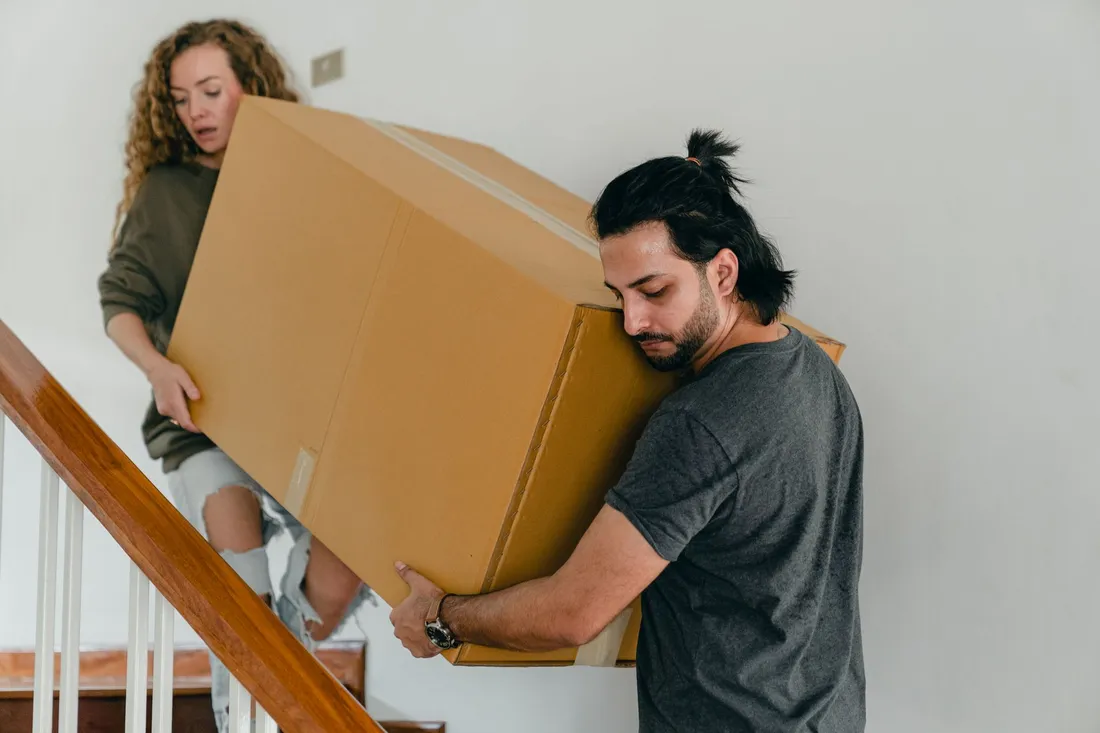 Zwei Umzugshelfer tragen einen großen unbeschrifteten Karton gemeinsam die Treppe hinunter